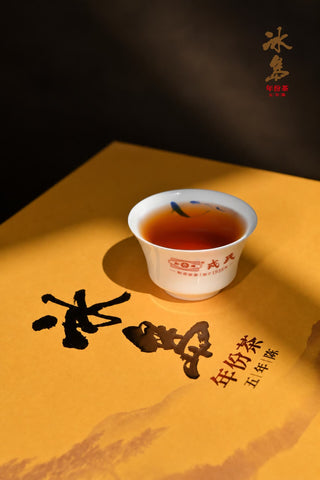 Small porcelain cup filled with Mengku Rongshi Bingdao Aged Ripe Pu-erh deep red liquor, sitting on the yellow gift box.