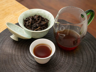 Brewed Mang Fei ripe pu-erh tea set with a celadon gaiwan, a filled white tasting cup, and a glass pitcher.