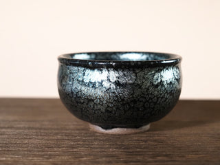Front low-angle view of a Jiang Jun Bei Jianzhan tea bowl with silver oil-spot pattern and broad low silhouette, on a wooden surface.