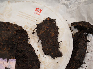 Chunks and loose dry tea leaves of 2017 Dayi 7452 ripe puerh tea pried open and resting on a paper wrapper.
