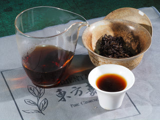 A complete Puerh tea tasting set featuring dry nuggets, a glass pitcher of tea, and a white tasting cup on an Orientaleaf tea mat.
