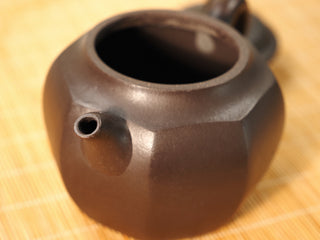 Top close-up of Shihuang Bafang Yixing teapot spout opening and sharp facet edges on dark clay body, bamboo mat