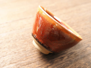 Tilted view of a Shu Kou red hare's-fur Jianzhan bowl on a wooden table, showing the dense dark streaks across the red-orange exterior and the small unglazed foot ring.