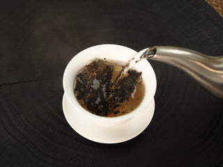 Boiling water poured over The Little Brick fu brick tea chunks in a white gaiwan, steam rising above the amber infusion