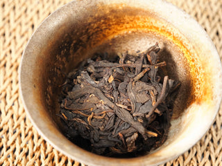 Dry leaves of Jing Chang Hao Quiet Years 2018 raw Pu-erh weighed in a rustic ceramic cup, dark twisted leaf with visible stems