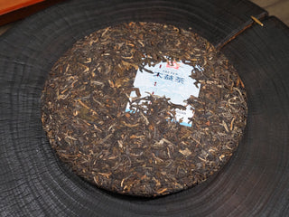 Whole tea cake of 2021 Dayi Mi Xiang Sheng Puerh tea resting flat on a dark wooden tea board.
