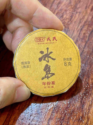 Fingers holding a wrapped 8g Mengku Rongshi Bingdao Aged Ripe Pu-erh coin cake, golden wrapper with calligraphy.