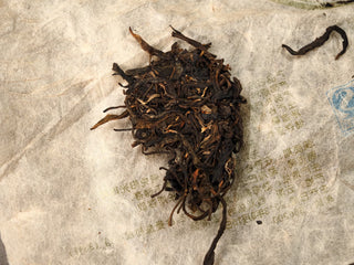 Broken-open chunk of Jing Chang Hao Mengsong Autumn Harvest 2013 raw Pu-erh cake revealing dark interior arbor leaves on the paper wrapper