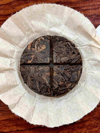 Unwrapped 8g Mengku Rongshi Bingdao Aged Ripe Pu-erh coin cake on rice paper, showing the cross-score for four quarters.