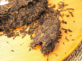 Broken edge of the Bojun Ripe Pu-erh 7.0 cake on a wooden tray, loose fermented leaves with golden buds scattered around.