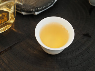 A single white porcelain tasting cup filled with bright, clear golden-apricot color white tea soup on a dark background.