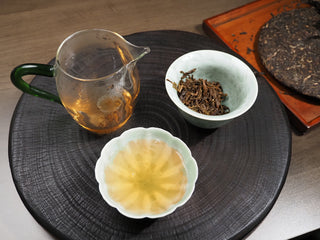 Gongfu tea ceremony set with 2021 Dayi Guanchao raw puerh tea soup in a porcelain cup, wet leaves in gaiwan, and glass fair cup.

