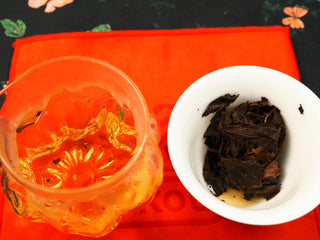 Glass fairness pitcher of amber tea liquor beside a gaiwan with steeped 2024 Shou Mei and Chen Pi waffle cake on red cloth.