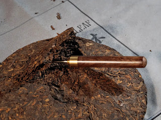 Bulang Mountain Ancient Tree Ripe Pu-erh tea cake on tea towel with a tea needle prying a small piece, demonstrating traditional handling