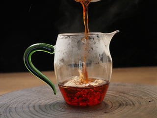 Clear and bright amber-red ripe puerh tea pouring from a Gaiwan into a glass fairness cup (Cha Hai) with a green handle.