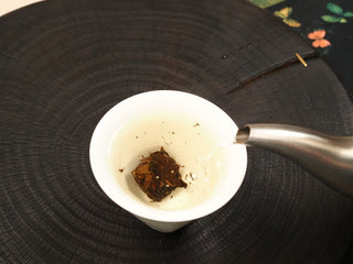 Hot water being poured from a metal kettle over 2014 Fuding White Peony waffle cake section in a cup, leaves beginning to infuse.