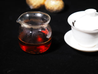Deep red, clear, and bright Chenpi Pu-erh tea liquor in a glass fairness cup next to a white gaiwan.