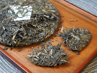 Flat lay display of 2025 Tan Chun raw Puerh tea cake alongside loose dry leaves on a wooden tray.