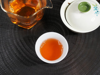 Bright orange-yellow tea soup of 2016 Dayi 7532 raw Puerh in a small white porcelain cup next to a glass server.