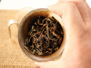 Wet leaves of Jing Chang Hao Quiet Years 2018 raw Pu-erh after steeping, opened brown-green arbor leaf held in a rustic ceramic cup