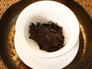 Dry Bojun Ripe Pu-erh 7.0 leaves loaded in a white porcelain gaiwan before brewing, on a dark metal saucer over a woven mat.