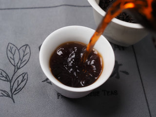 Pouring rich red Dayi 7572 ripe puerh tea from a fairness cup into a white porcelain teacup.
