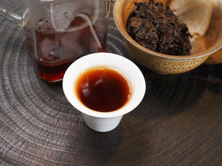 A small white porcelain cup filled with rich, dark burgundy-red Dayi 7452 ripe puerh tea liquor.