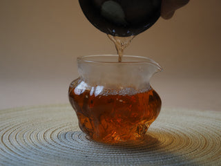 Pouring clear, bright amber-gold brewed tea liquor of the 2005 Lincang vintage raw Pu-erh into a glass pitcher.