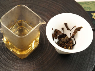 Glass fairness pitcher of amber liquor beside a white ceramic gaiwan with steeped 2014 White Peony waffle cake leaves.