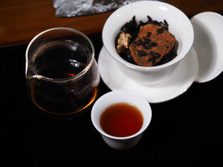 Chinese Gongfu tea setup showcasing a cup of dark red citrus Pu-erh tea, glass pitcher, and gaiwan.
