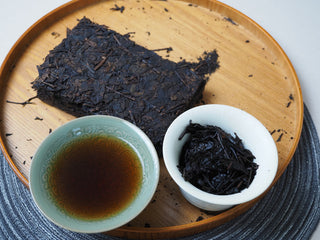 Complete tea presentation showing the tea brick on a wooden tray alongside a cup of dark red tea liquor and brewed leaves.