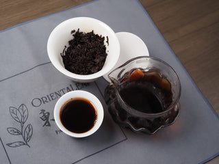 Chinese gongfu tea set with brewed Dayi 7572 ripe puerh tea, gaiwan, glass pitcher, and teacup on a mat.
