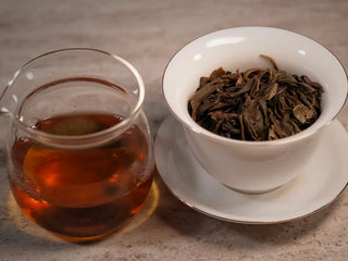 Gaiwan and glass pitcher side by side with 2019 Lincang Ancient Arbor Sheng Pu-erh Tea Cake showing bright yellow-green to reddish liquor and open leaf base
