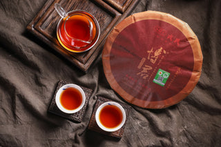 Overhead scene of 2019 Bojun Ripe Pu-erh: 500g cake, glass pitcher of red liquor, and two small cups of tea on a dark background.