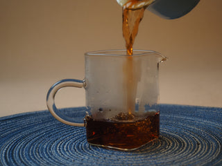 Pouring freshly brewed dark red Shou Pu-erh tea liquor into a glass fairness cup (cha hai)