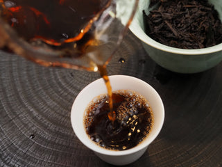 Pouring rich, sweet burgundy-red Dayi 7592 ripe puerh tea into a white-lined celadon teacup.