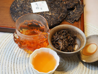 Complete tea brewing session setup featuring the 2005 Lincang vintage Sheng Pu-erh tea cake, liquor pitcher, and tasting cup.