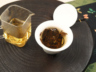 Open white gaiwan with dark steeped leaves of 2014 Fuding White Peony waffle cake beside a pitcher of amber tea liquor.