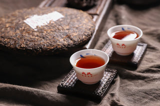 2019 Bojun 500g cake beside two small cups of red tea on wooden coasters, dark background with a linen cloth.