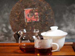 Qilin Hao Fengshan Gushu Ripe Pu-erh Tea Cake 2021 as a blurred background with Gaiwan, Gongdao cup, and tea cup in focus, highlighting the deep red tea liquor