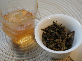 Bright and clear brewed tea liquor of 2026 Tan Chun raw Puerh served in a white tasting cup alongside a fairness pitcher.