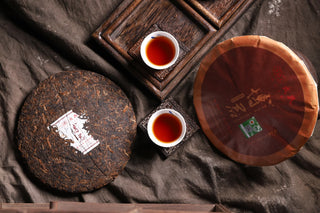 Overhead scene: two 2019 Bojun cakes (one unwrapped, one wrapped) and two small cups of red tea on wooden coasters.