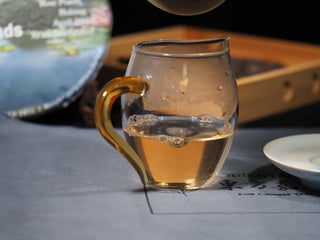 Bright, clean, and golden raw Puerh tea liquor in a glass pitcher with the Bulang Buds cake in the background.