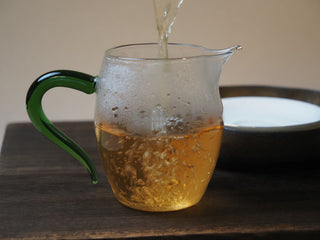 Pouring golden raw pu-erh tea liquor from gaiwan into glass fairness pitcher
