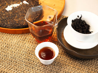 Gongfu brewing scene of Bojun Ripe Pu-erh 7.0: cake, glass pitcher of red liquor, tasting cup, and infused leaves in a gaiwan.