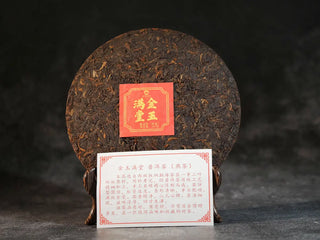 Golden Treasure Ripe Puerh Tea Cake – Menghai Origin, front view of the unwrapped tea cake revealing its dark brown leaves with golden tips, accompanied by a product information card placed below.