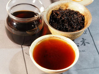 Golden Treasure Ripe Puerh Tea Cake – Menghai Origin, open gaiwan with glossy dark brown tea leaves, alongside a fairness cup and a teacup filled with deep reddish-brown brewed tea.