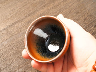 Handheld angled view of a Shu Kou hare's-fur Jianzhan bowl in palm, showing the contracted-mouth silhouette from a slight tilt, dark brown-to-black streaks across the interior, and a deep black center.