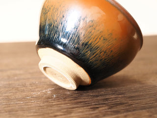 Low-angle side view of a Shu Kou hare's-fur Jianzhan bowl resting on its rim on a wooden table, showing the dense dark brown-to-black streaks on the exterior curving down to the unglazed foot ring.