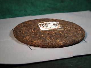 Spring Ming Ripe Pu-erh Tea Cake (2008 Commemoration) – Menghai, Yunnan resting on a tea towel, prepared for breaking and brewing, highlighting traditional tea setup.